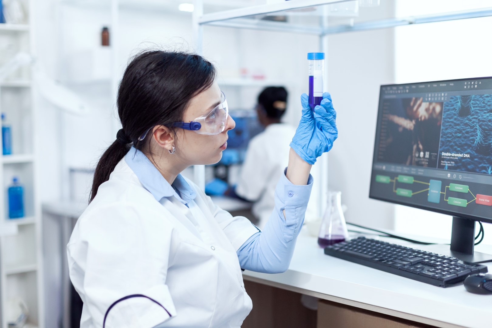 A woman in a lab looking at a test tube. She is sitting at a desk in front of a computer running tets.