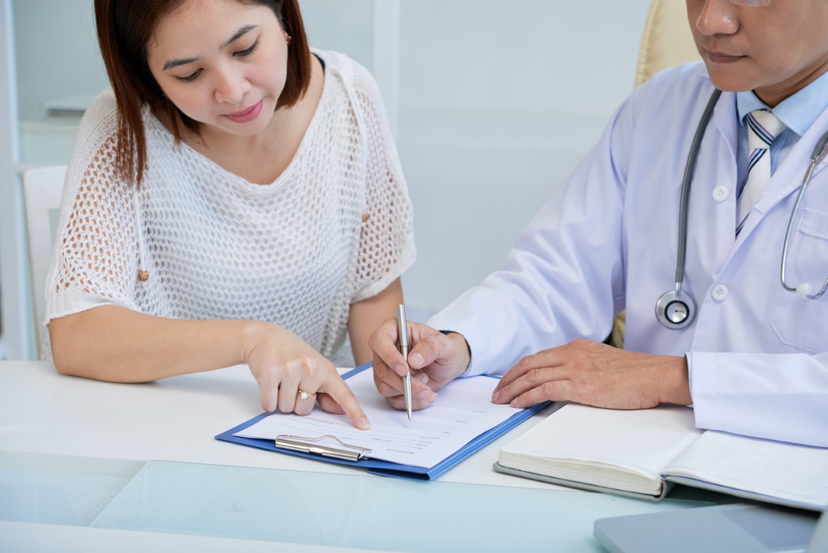 A woman and a male doctor sitting next to each other at a desk filling out paperwork on a clipboard.